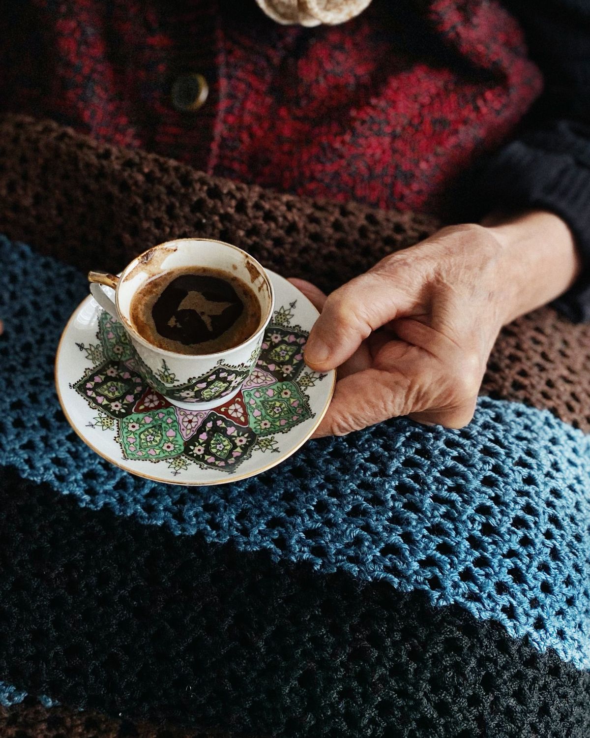 Eine ältere Hand hält eine Tasse Kaffee auf einer gehäkelten Decke.